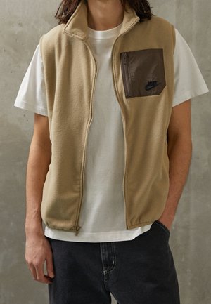 Person iført beige fleecevest med brun lynlåslomme på brystet over hvid t-shirt og mørke bukser, stående mod en grå tekstureret væg.