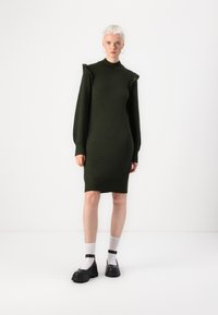 Robe tricotée verte à col montant, avec des accents d'épaule froncés et des manches longues, associée à des chaussures noires à semelles épaisses et des chaussettes blanches.