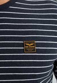 T-shirt rayé bleu marine avec des rayures horizontales blanches. Il présente un badge carré jaune et noir avec l'étiquette "PME LEGEND" sur la poitrine.