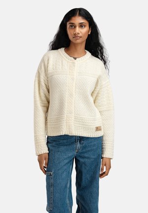 Kvinne med langt svart hår iført en kremfarget strikkegenser og blå jeans, stående mot en hvit bakgrunn.