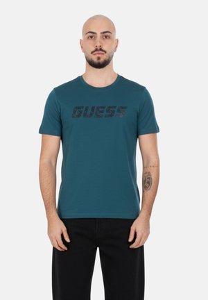 Uomo con barba e tatuaggio sull'avambraccio sinistro, indossa una maglietta a maniche corte color turchese con la scritta "GUESS", in piedi contro uno sfondo bianco.