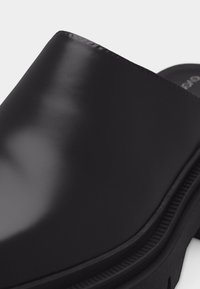 Chaussure noire en cuir lisse à enfiler, avec une semelle épaisse et un bout arrondi, présentée sur un fond clair uni.