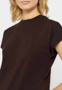 Γυναίκα με ξανθά μαλλιά που φοράει καφέ σκούρο κοντομάνικο τοπ με στρογγυλή λαιμόκοψη, μπροστά σε απλό φωτεινό φόντο.