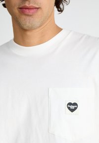 White cotton t-shirt featuring a breathable crew neck and a square pocket with a black heart logo label. Smooth texture, relaxed fit.