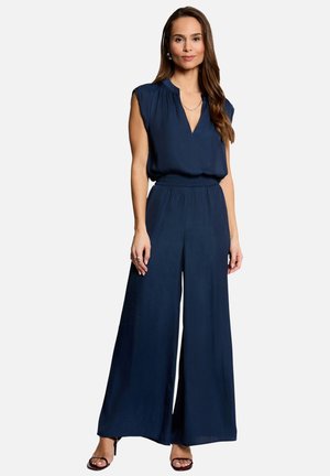 Femme debout portant un chemisier sans manches bleu marine avec un décolleté en V et un pantalon à jambes larges, avec des sandales, sur un fond clair uni.