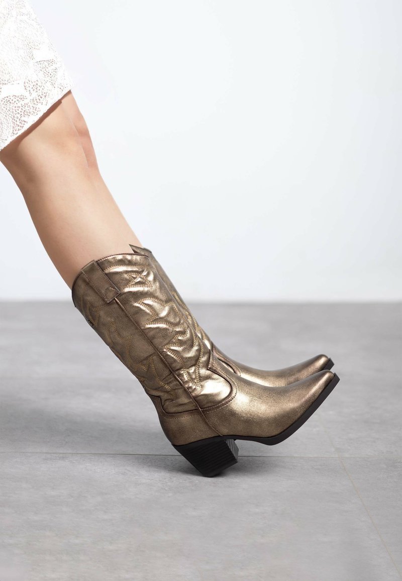Botas de cowboy metálicas doradas con punta afilada y tacón cuadrado, que presentan detalles de costura texturizada a lo largo del eje.