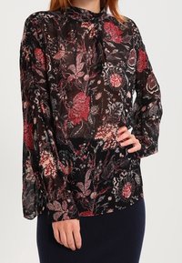 Blusa nera trasparente con motivo floreale in rosso, rosa e crema, caratterizzata da maniche lunghe e un collo alto con dettaglio a nastrino.