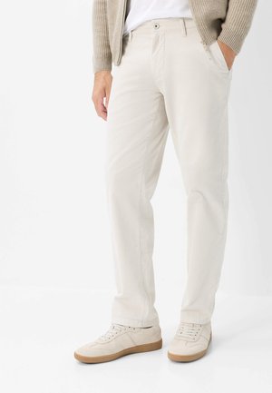 Mand iført beige casual bukser, beige sneakers, hvid skjorte og beige strikket jakke, stående med den ene hånd i lommen.