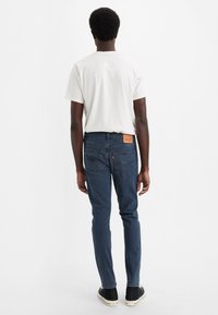 Tamnoplave traperice uskog kroja s džepovima na leđima i vidljivom kožnom oznakom brenda. Kombinirane s običnom bijelom majicom.