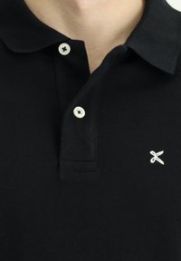 Camisa polo negra con cuello clásico, dos botones y un pequeño detalle bordado en blanco en el lado izquierdo. Tejido de algodón con una textura suave.