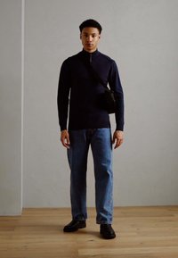 Suéter de cremallera azul marino, jeans de mezclilla claros y zapatos negros, usados por una persona de pie contra un fondo neutro. Pequeño bolso negro cruzado.