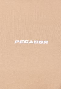 Il tessuto a costine color tan presenta la scritta "PEGADOR" in bianco. Il design è liscio e testurizzato, offrendo un aspetto semplice e moderno.