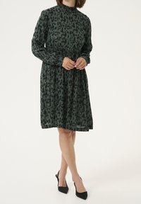 Grünes Kleid mit schwarzen abstrakten Druck, hoher Ausschnitt, lange Ärmel und ein ausgestellter Rock. Kombiniert mit schwarzen Absatzschuhen für das Styling.