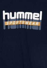 Camiseta azul marino con un logo "hummel SPORTSWEAR" en blanco y naranja, con un diseño texturizado detrás del texto. Fabricada con un tejido suave.