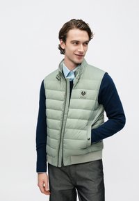 Lysegrøn polstret vest med lynlås foran, høj krave, sidelommer og et logo-patch. Båret over en navyblå sweater og kombineret med grå bukser.