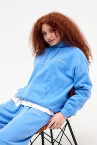 Junge Frau mit lockigem roten Haar trägt einen blauen Kapuzenpullover und Sweatpants und sitzt auf einem Stuhl mit Metallrahmen vor einem weißen Hintergrund.