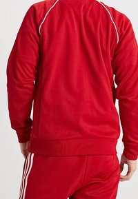 Punainen urheilu takki, joka on valmistettu sileästä kankaasta ja jossa on valkoiset saumat hartioiden kohdalla sekä ribbaus vyötäröllä.