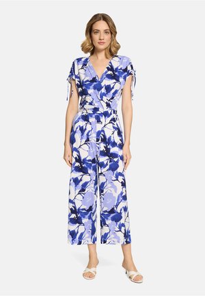Kvinde iført en ærmeløs, slå-om-style jumpsuit med blå og hvide blomsterprint og hvide hælede sandaler, stående mod en ensfarvet baggrund.
