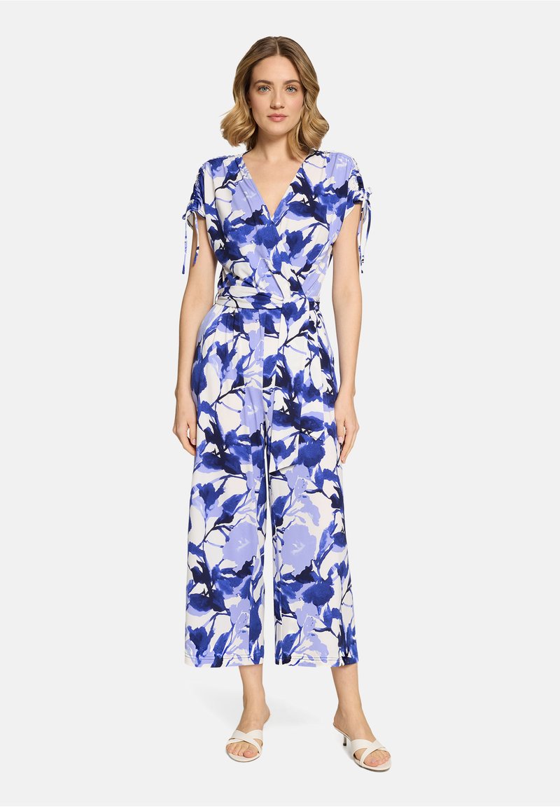 Femme portant une combinaison sans manches de style portefeuille avec un imprimé floral bleu et blanc et des sandales blanches à talons, se tenant devant un fond uni.