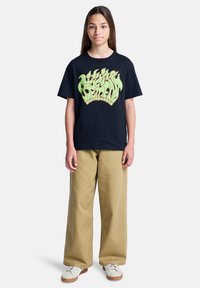 Έφηβη κοπέλα με μακριά καστανά μαλλιά, φορώντας μαύρη μπλούζα με σχέδιο, φαρδιά μπεζ παντελόνια και λευκά αθλητικά παπούτσια, που στέκεται μπροστά σε λευκό φόντο.