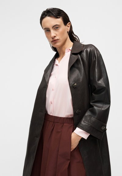 Καφέ δερμάτινο παλτό τρανς με μεγάλα κουμπιά, πάνω από ροζ ανοιχτό πουκάμισο, σε συνδυασμό με πλισέ μπορντώ φούστα. Ομοιόμορφη υφή σε όλο το ένδυμα.