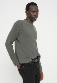 Νεαρός άνδρας που φοράει γκρι πλεκτό πουλόβερ και μαύρα τζιν με μαύρη ζώνη, ποζάροντας με το ένα χέρι στην τσέπη, μπροστά σε λευκό φόντο.