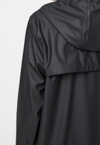 Veste noire imperméable avec capuche, présentant une texture lisse et brillante ainsi qu'un design avec empiècement au dos. Lignes épurées et style minimaliste.