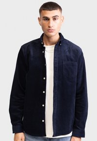 Chemise en velours côtelé bleu marine avec fermeture à boutons, poche poitrine unique et col classique. Portée sur un t-shirt à col rond de couleur claire.