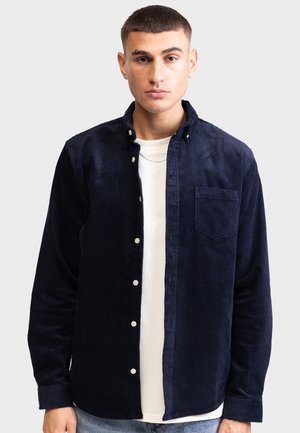 Marineblauwe corduroy shirt met een button-down kraag, een voorzak en twee knopen, gedragen over een wit T-shirt. Heeft een gemêleerde stof en lange mouwen.
