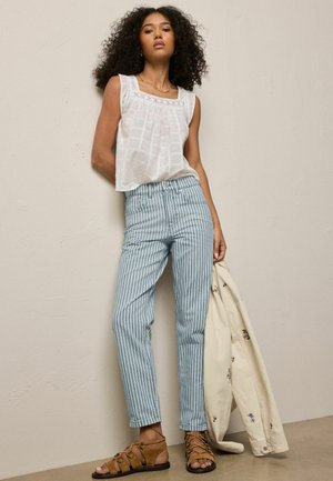 Frau mit lockigem Haar lehnt an der Wand, trägt ein ärmelloses weißes Oberteil, blau-weiß gestreifte Hose, braune Sandalen und hält eine cremefarbene Jacke mit Blumenstickerei.