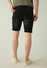 Shorts en denim noir avec un ourlet retroussé, dotés de poches arrière et d'une texture délavée, associés à des baskets beiges sur un fond clair.