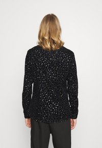 Černý velurový top se vzorem zvířecí tisku, zdobený zlatými a stříbrnými flitry. Dlouhé rukávy a zaoblený lem dodávají uvolněný střih.