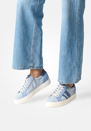 Person trägt hellblaue Jeans und hellblaue Sneaker mit weißen Sohlen, Schnürsenkeln und einem Reißverschlussdetail an der Seite.
