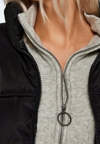 Svart vadderad väst med dragkedja fram, bärs över en ljusgrå fleece-tröja. Noterbara runda dragkedje-hanter och texturerade tygdetaljer.