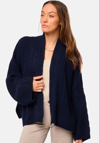 Cardigan tricoté bleu marine avec un design en cable texturé, manches larges, devant ouvert et accents côtelés. Tissu léger et confortable.