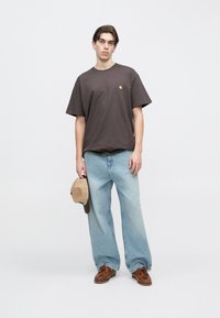 Νεαρός άνδρας στέκεται φορώντας ένα σκούρο φαρδύ μπλουζάκι, ανοιχτό μπλε τζιν παντελόνι με φαρδιά μπατζάκια, καφέ παπούτσια και κρατώντας δίπλα του ένα καφέ καπέλο.