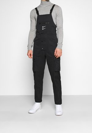 Czarne ogrodniczki z regulowanymi szelkami, suwakiem na kieszeni na piersi oraz bocznymi kieszeniami cargo. Zestawione z szarą, długorękawową koszulką z golfem i białymi sneakersami.