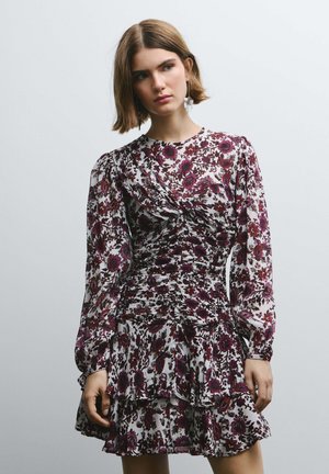 Vestido floral con mangas largas, corpiño fruncido y dobladillo con volantes en capas. Presenta un fondo blanco con patrones florales en púrpura y burdeos.