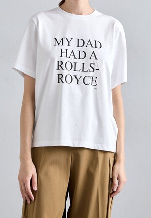 Osoba ubrana w białą koszulkę z czarnym napisem „MÓJ TATA MIAŁ ROLLS-ROYCE” oraz brązowe spodnie na jednolitym tle.