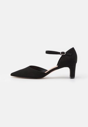 Black suede pump with a pointed toe, ankle strap featuring a circular buckle, and a low block heel. Smooth texture and minimalist design.