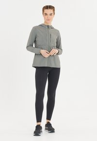 Lätt, grå zip-jacka med huva, långa ärmar och en lätt hög-låg fåll, ihop med svarta leggings och svarta träningsskor.