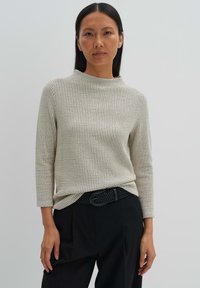 Cremfarbener Waffelstricktop mit höherem Kragen und Dreiviertelärmeln, kombiniert mit hochgeschnittenen schwarzen Hosen und einem geflochtenen schwarzen Gürtel.