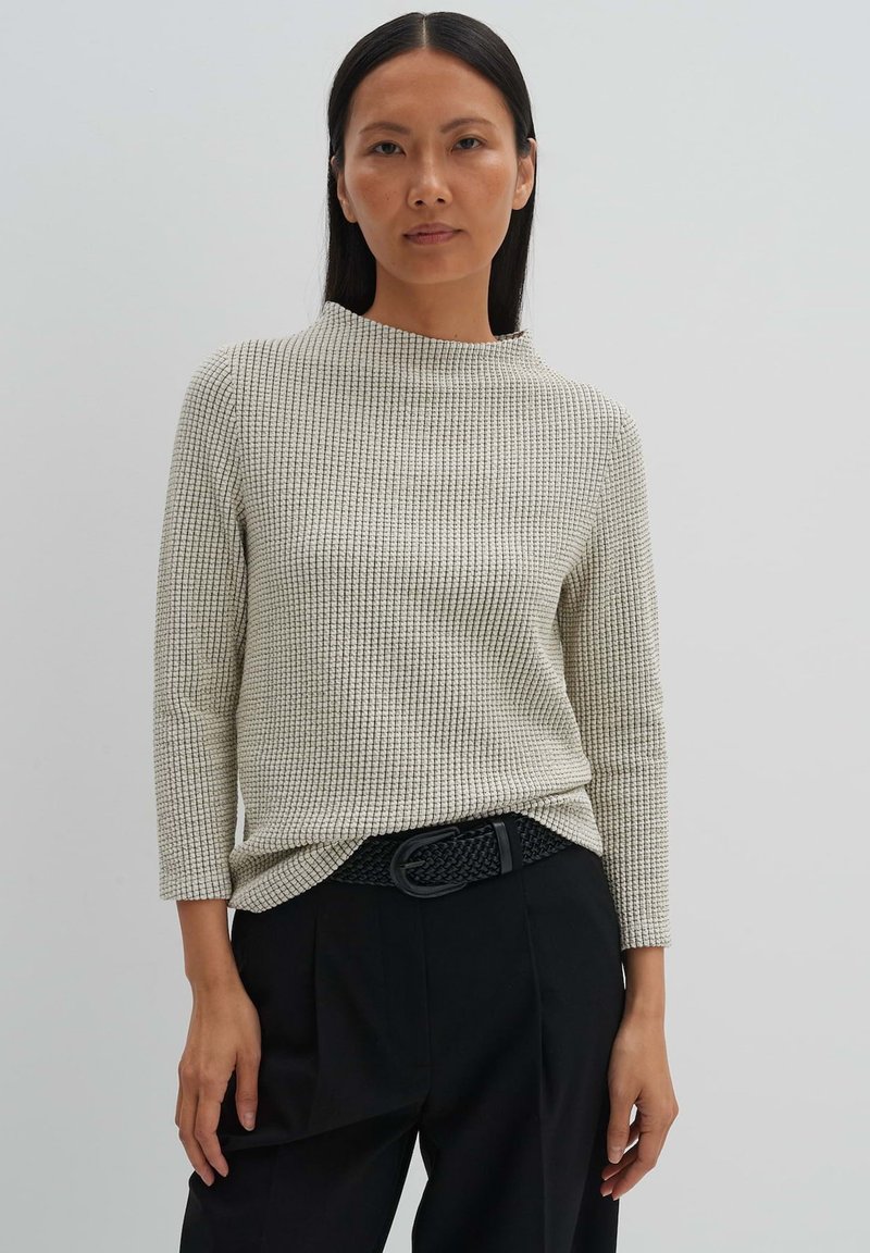 Cremfarbener Waffelstricktop mit höherem Kragen und Dreiviertelärmeln, kombiniert mit hochgeschnittenen schwarzen Hosen und einem geflochtenen schwarzen Gürtel.