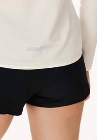 Weißes Langarmoberteil mit einem strukturierten Muster und subtiler grauer Grafik am unteren Ende, kombiniert mit schwarzen Sportshorts.
