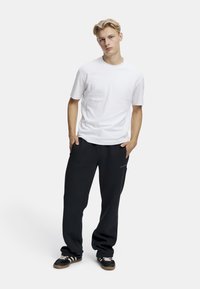 Vit bomull t-shirt i kombination med svarta träningsbyxor, med sidofickor och en liten textlogga på vänster lår. Svarta sportskor.