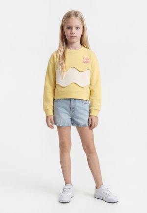 Ξανθιά κοπέλα στέκεται με το πρόσωπο προς τα εμπρός, φοράει κίτρινο φούτερ με την επιγραφή «Club Soleil», ανοιχτόχρωμα τζιν σορτς και λευκά αθλητικά παπούτσια σε λευκό φόντο.