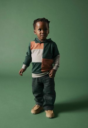 Felpa color-block verde e arancione con il testo "SII FANTASTICO", abbinata a jeans scuri e scarpe beige, su uno sfondo verde.