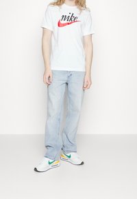 Valkoinen lyhythihainen T-paita, jossa on musta ja punainen "Nike"-logo, yhdistettynä vaaleansinisiin farkkuihin ja valkoisiin lenkkareihin, joissa on vihreitä ja keltaisia yksityiskohtia.