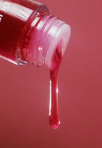 Bottiglia di vetro trasparente con tappo bianco, che versa un liquido lucido di colore rosa intenso che gocciola su uno sfondo rosa abbinato.
