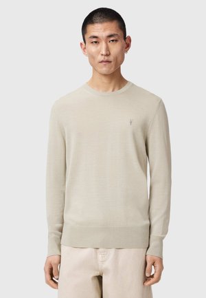 Mężczyzna o krótkich włosach, ubrany w jasnobeżowy sweter z długim rękawem i pasujące jasne spodnie, stojący na tle jednolitego tła.